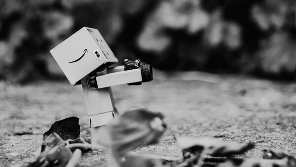 White box character with Amazon smile logo tilting head while holding camera on rocky ground.