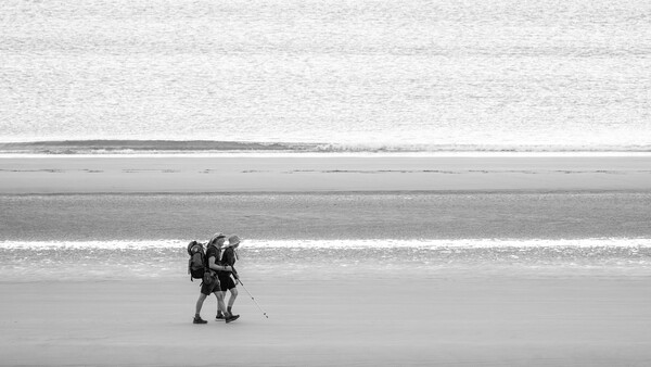 Two figures walking together on a vast, minimalist beach with horizontal bands of sand and water.