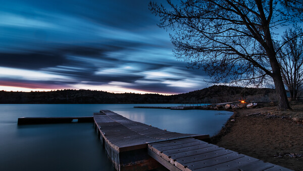 Wooden dock extending into a still lake at dusk, with bare trees and distant shoreline beneath a dramatic twilight sky.