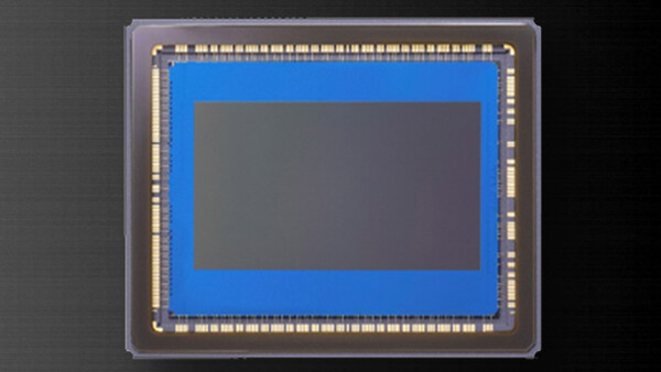 Close-up photograph of a camera image sensor die mounted in a clear plastic package with gold contact pins around the perimeter.