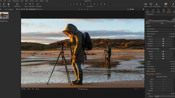 Photographer in yellow jacket operating tripod-mounted camera on beach at golden hour with two figures in distance.