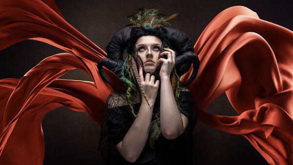 Person in dark costume with feathered headdress and dramatic red fabric wings against black background.