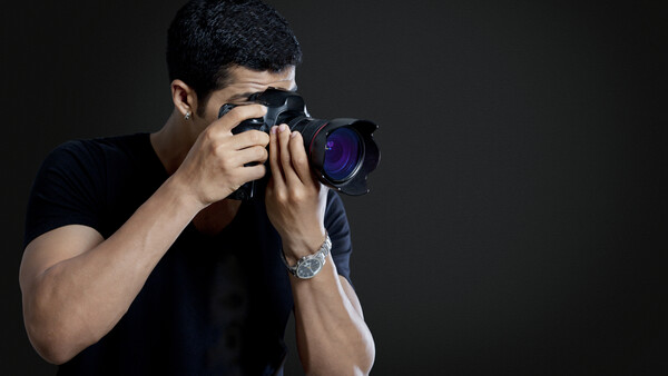 Male photographer holding a DSLR camera with telephoto lens, positioned against dark background.
