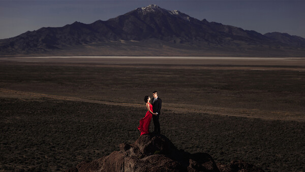 Couple in formal attire standing on a dark rock formation in an expansive desert plain, with a snow-capped volcanic mountain in the distance.