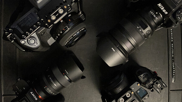 Professional camera gear layout including Nikon bodies, lenses with hoods, and accessories arranged on a dark surface.