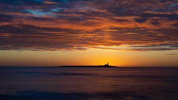 Silhouetted lighthouse on a distant island at sunset over calm ocean waters.