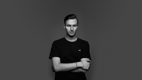 Young man in black t-shirt with arms crossed, looking at camera against gray background.