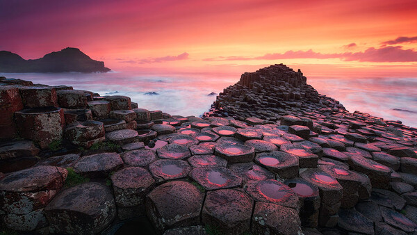 Hexagonal basalt columns in foreground with golden sunset sky and rocky islands in background.