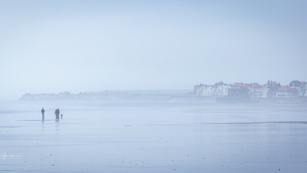 Two figures walking on a misty, wet beach at dawn with a coastal town silhouetted in the distance.