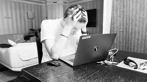 Person in white shirt holding head while working at laptop in hotel room.