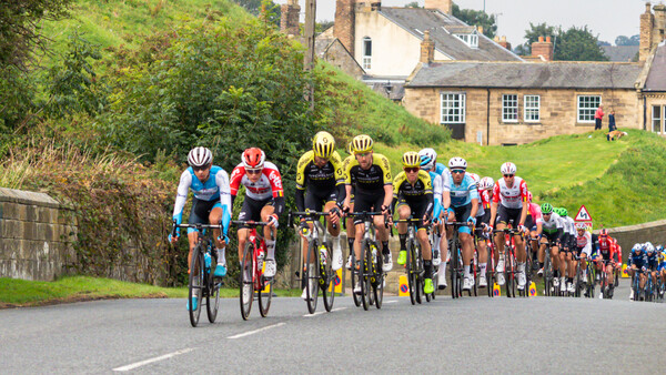 Professional cycling race with peloton riding on rural road past stone buildings and green hillsides.