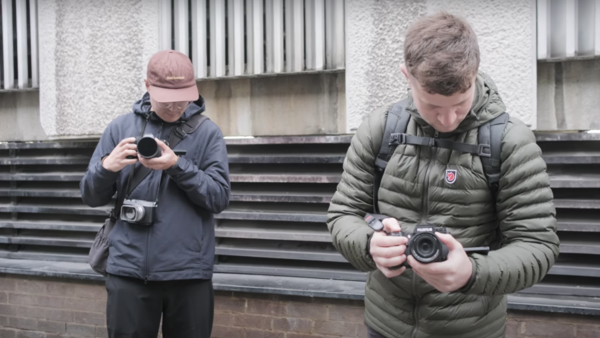 Two photographers holding compact cameras outside a modern building with corrugated metal siding.