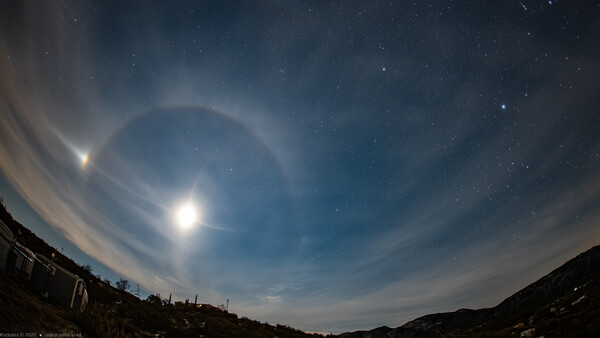 Bright moon with concentric halo rings in twilight sky above dark mountain silhouettes.