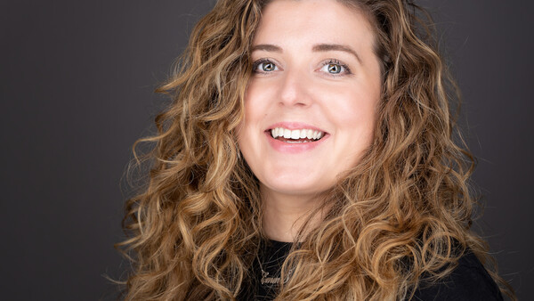 Woman with long wavy brown hair smiling at camera against dark background.
