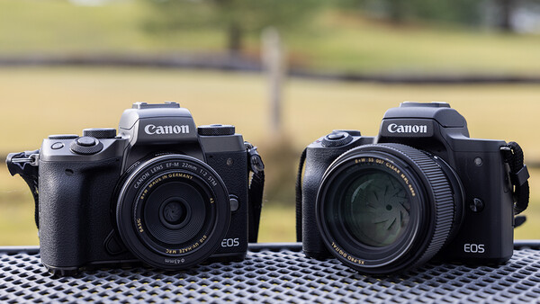 Two Canon EOS mirrorless cameras side by side on a checkered surface with blurred green background.
