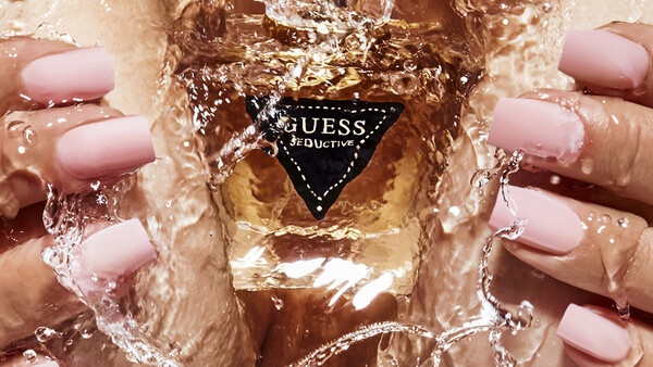 Hands with pink nail polish holding a GUESS fragrance bottle submerged in water.