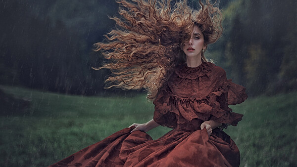 Woman in burgundy dress with windswept long hair in heavy rain against dark green field.