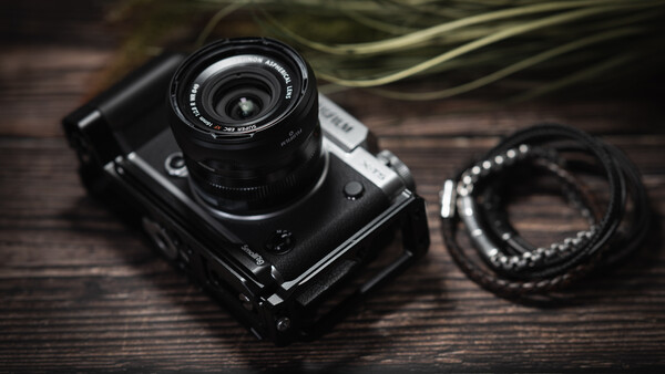 Black rangefinder camera with lens on wooden surface next to beaded bracelet.