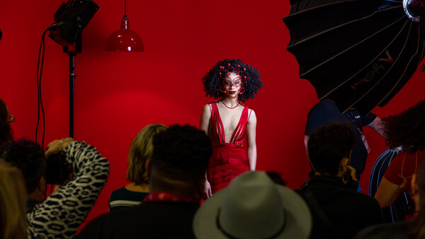 Woman in red dress posed against vibrant red backdrop during studio photoshoot with visible lighting equipment.
