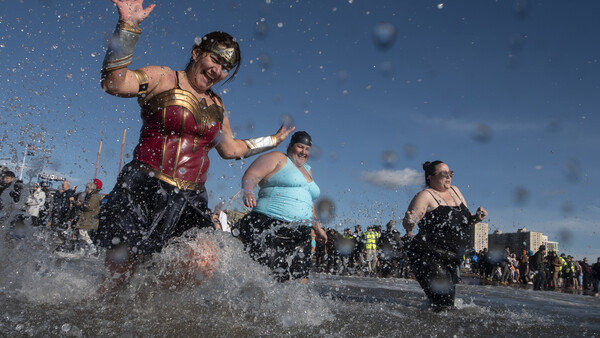 Multiple people running through shallow water on a beach under blue sky with water splashing around them.