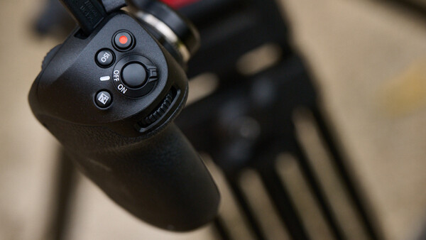 Close-up of a camera gimbal's control handle with buttons and a rotary dial in sharp focus, mounted on a tripod in the blurred background.
