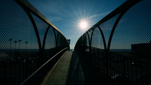 Silhouetted pedestrian bridge with mesh railings framing a bright sun against blue sky.