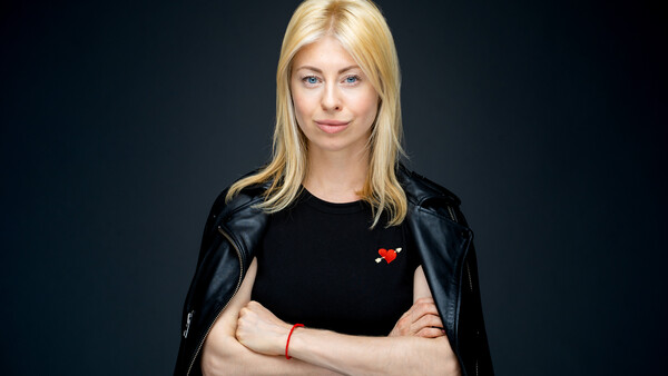Blonde woman in black leather jacket and black shirt against dark background, arms crossed.