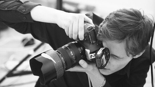 Photographer holding a DSLR camera with telephoto lens up to their face, shooting in black and white.