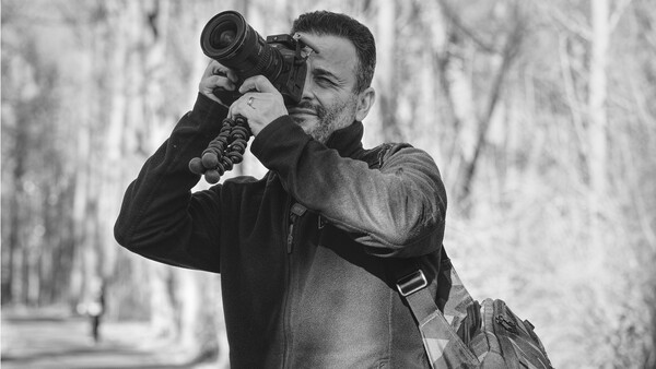 Photographer holding a telephoto lens camera to his eye while standing outdoors among bare trees.