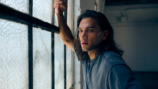 Man with long dark hair gazing toward a barred window with strong directional side lighting.
