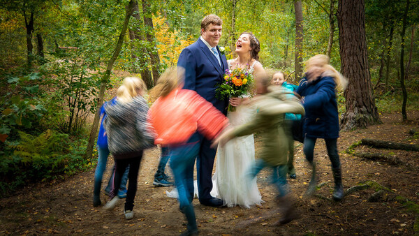 Bride and groom surrounded by blurred children running in a forest clearing during a wedding celebration.