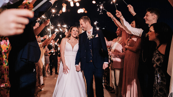 Bride and groom walk through a tunnel of sparklers held by wedding guests at night.