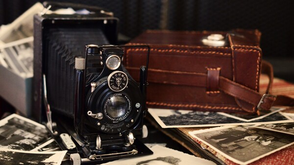 Vintage folding camera displayed with leather case, scattered black and white photographs, and another camera in soft studio lighting.