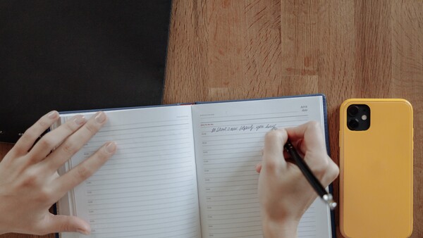 Person's hands writing in an open notebook with a pen, with a yellow smartphone visible on a wooden desk.