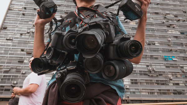 Photographer holding multiple Canon cameras and lenses against urban building backdrop.