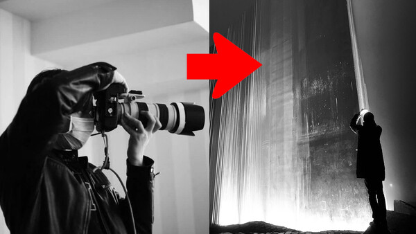 Photographer holding a camera with a telephoto lens while being photographed by another person in a minimalist interior space.