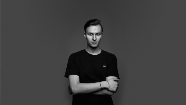 Young man in black t-shirt with arms crossed, standing against neutral gray background.