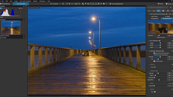 Long wooden pier at dusk with street lamps reflecting on wet surface under blue twilight sky.