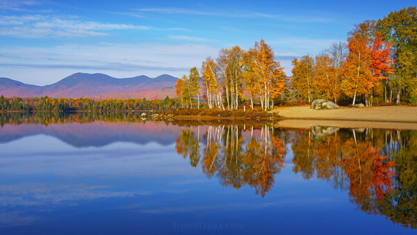 Autumn forest reflected in still lake water with mountains in the background.