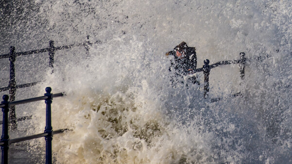 Person standing on a metal railing as a large wave crashes over them with intense spray and foam.