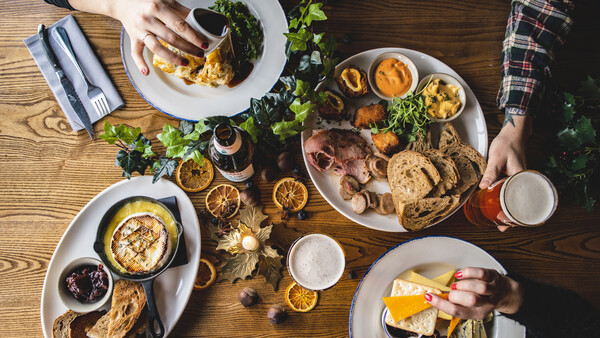 Overhead view of a shared meal with multiple white plates of food, fresh herbs, and beverages on a wooden table.