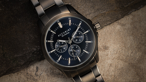 Black stainless steel chronograph watch with three subdials on a textured concrete surface.