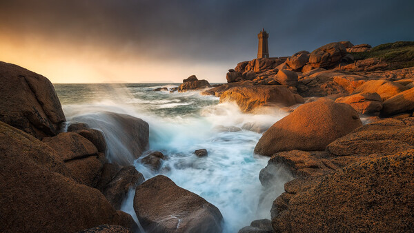 Lighthouse on rocky coastline at golden hour with waves crashing against granite boulders.