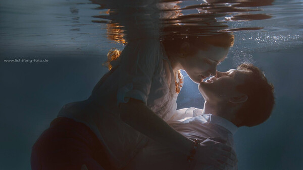 Two people embracing underwater in shallow water with natural sunlight filtering from above.