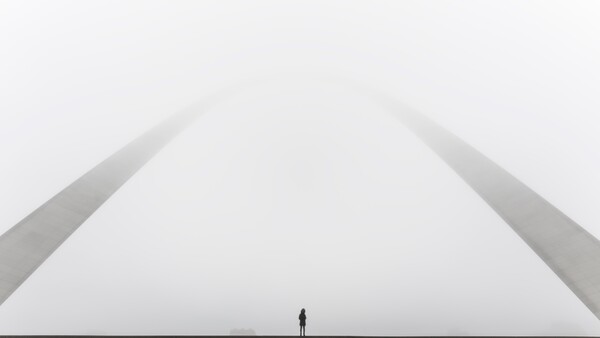 Silhouette of a solitary figure standing beneath a monumental curved architectural structure against an overcast sky.
