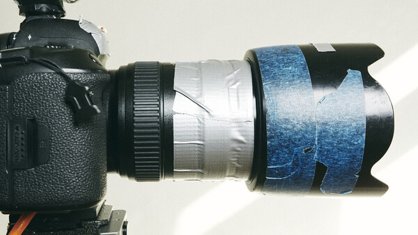 DSLR camera with lens wrapped in blue gaffer tape and clear tape showing wear and damage.