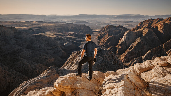 Hiker standing on rocky outcrop overlooking expansive canyon landscape with layered mountains.
