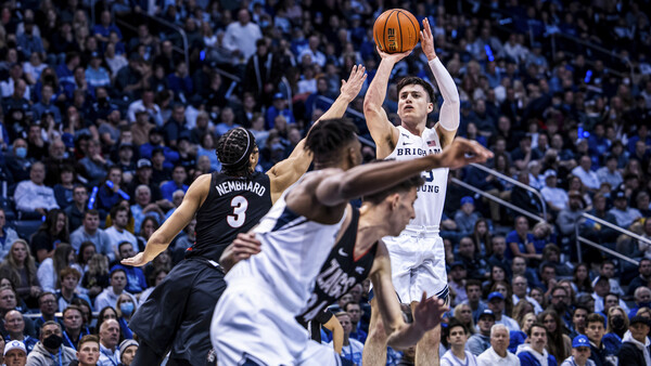 Basketball player in white jersey shooting over defender in black jersey during game with crowd in background.