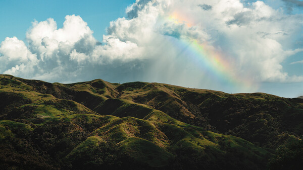 Rolling green hills beneath a blue sky with white clouds and a rainbow in the distance.