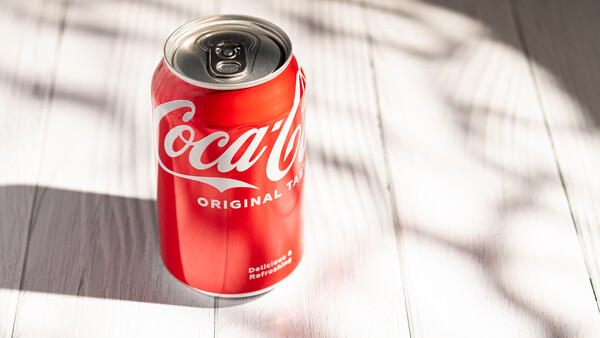 Red Coca-Cola Original can with pull-tab top on white wooden surface with dappled shadow.